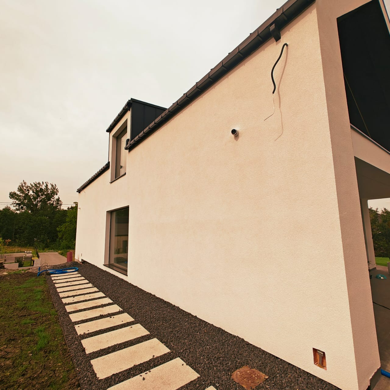 Nowoczesny dom z jasną, ziarnistą elewacją, czarnym dachem i ścieżką z płyt. Widoczna kamera, wystające kable i niedokończone otoczenie.