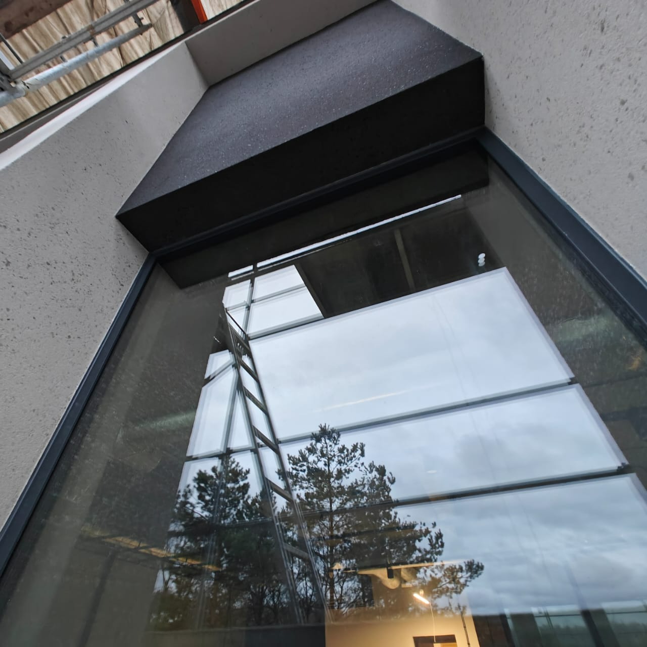 Szczegół okna z czarną ramą i ociepleniem nad nim. W szybie odbija się drabina i drzewo. Widoczny fragment elewacji w jasnym odcieniu.