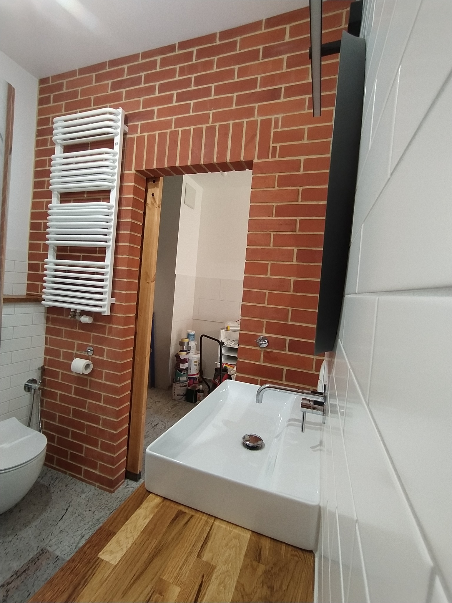 Nowoczesna łazienka z ceglaną ścianą, białymi płytkami i drewnianym blatem pod umywalką, widoczny grzejnik drabinkowy i fragment toalety. W tle widoczne farby i narzędzia.