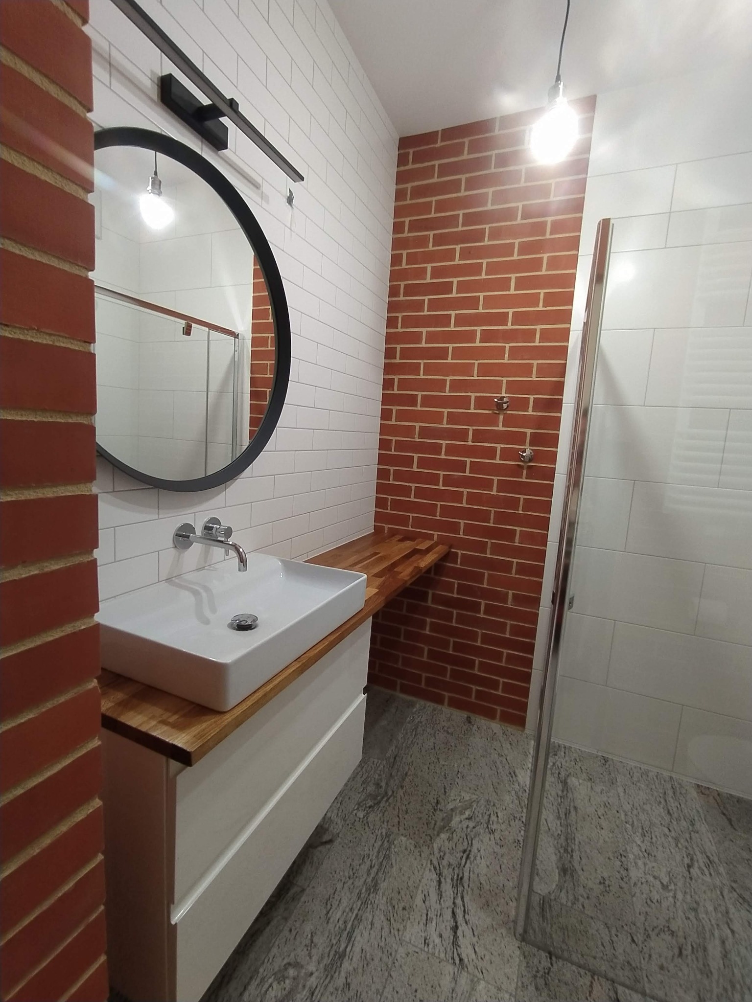 Nowoczesna łazienka z białymi płytkami, ścianą z czerwonej cegły, okrągłym lustrem z czarną ramą, białą umywalką nablatową na szafce z drewnianym blatem i szarą podłogą z granitu.