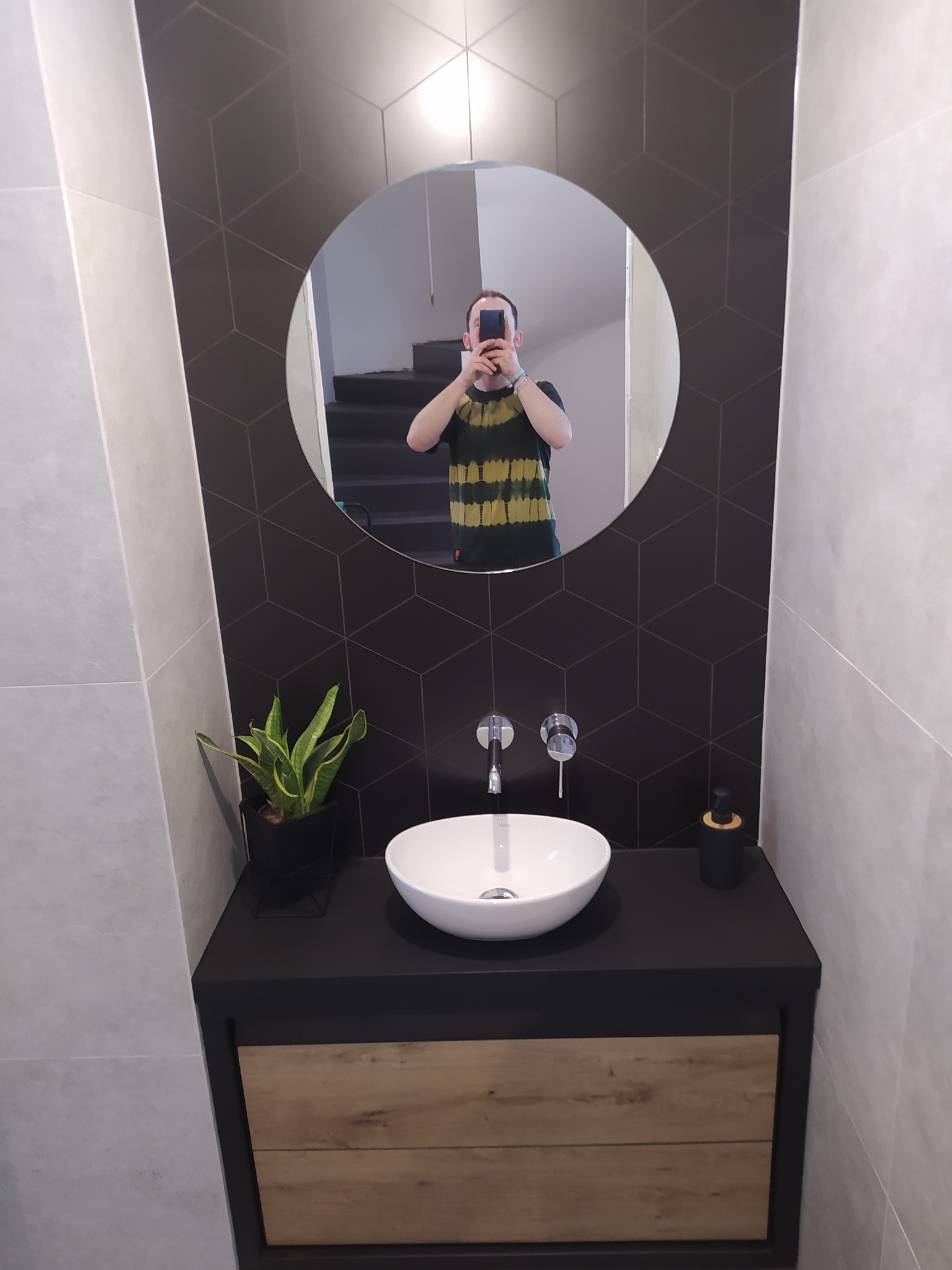 Nowoczesna łazienka z umywalką nablatową, czarnym blatem, drewnianą szufladą i geometryczną mozaiką na ścianie, odbicie fotografa w okrągłym lustrze.