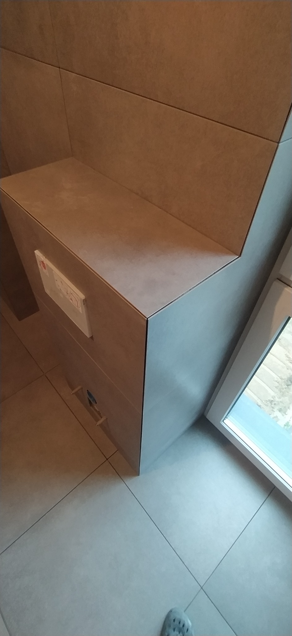 Szara, ceramiczna obudowa stelaża WC podtynkowego z widocznym przyciskiem spłukującym, dopasowana kolorystycznie do płytek na ścianie i podłodze w łazience.