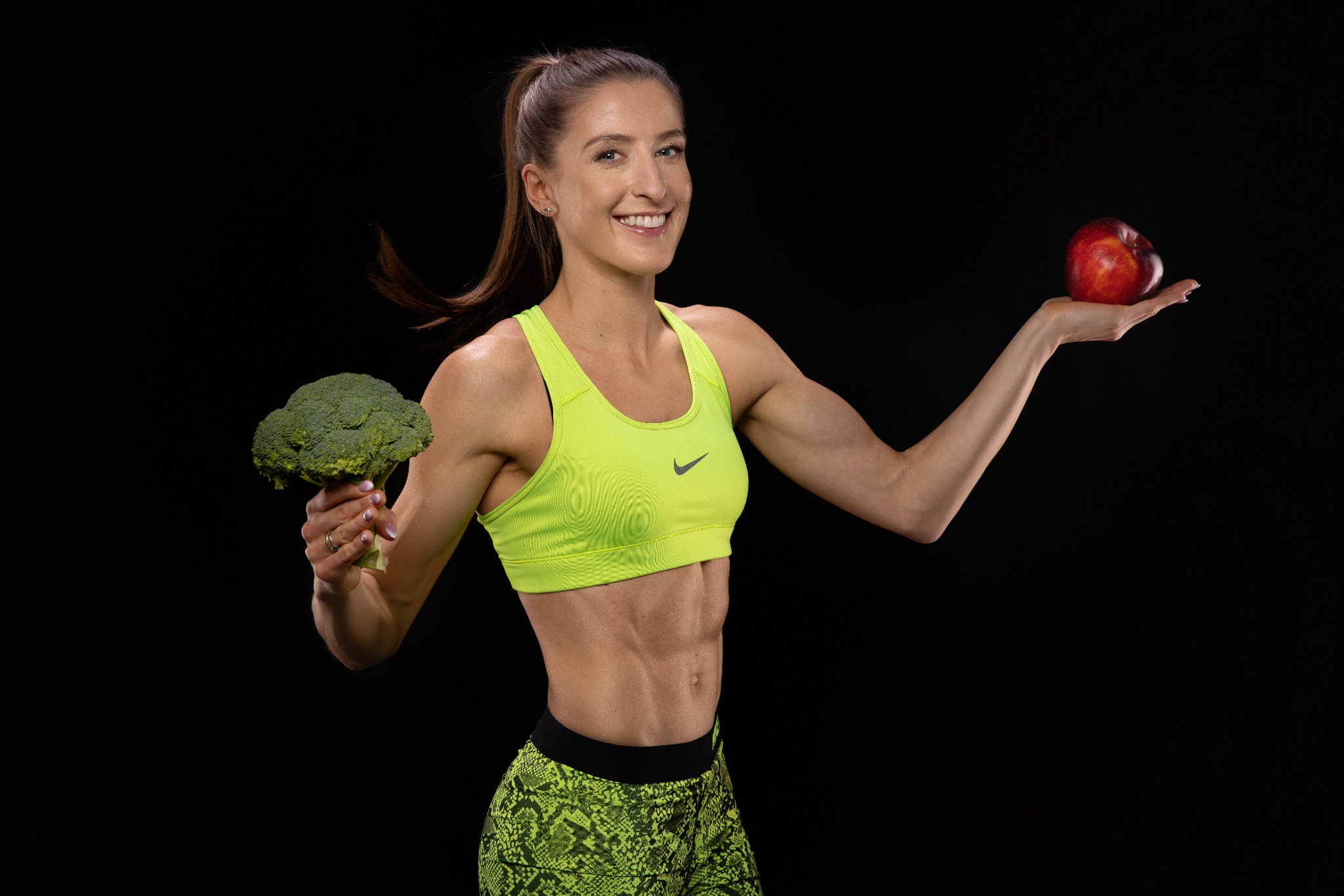 Uśmiechnięta, wysportowana kobieta w neonowym stroju sportowym prezentuje brokuł w jednej dłoni i nektarynkę w drugiej na czarnym tle.