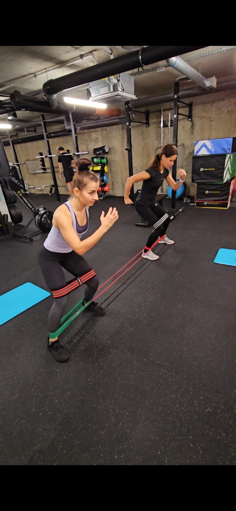 Dwie kobiety ćwiczą w siłowni z gumami oporowymi na nogach, wykonując ćwiczenia wzmacniające, widoczny sprzęt do ćwiczeń siłowych w tle.