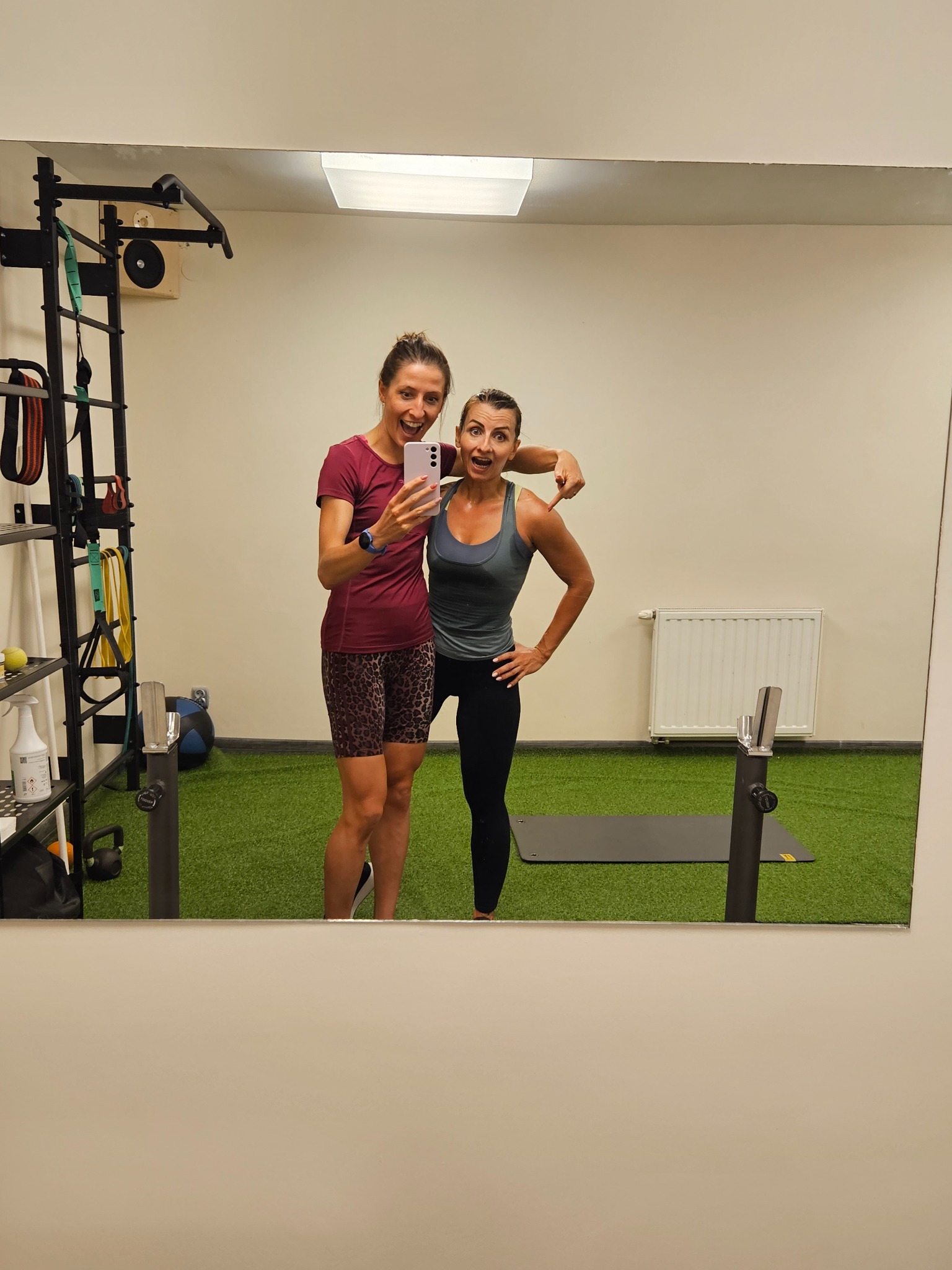 Dwie uśmiechnięte kobiety robią selfie w lustrze na tle siłowni z drabinkami, sztuczną trawą i matą do ćwiczeń.
