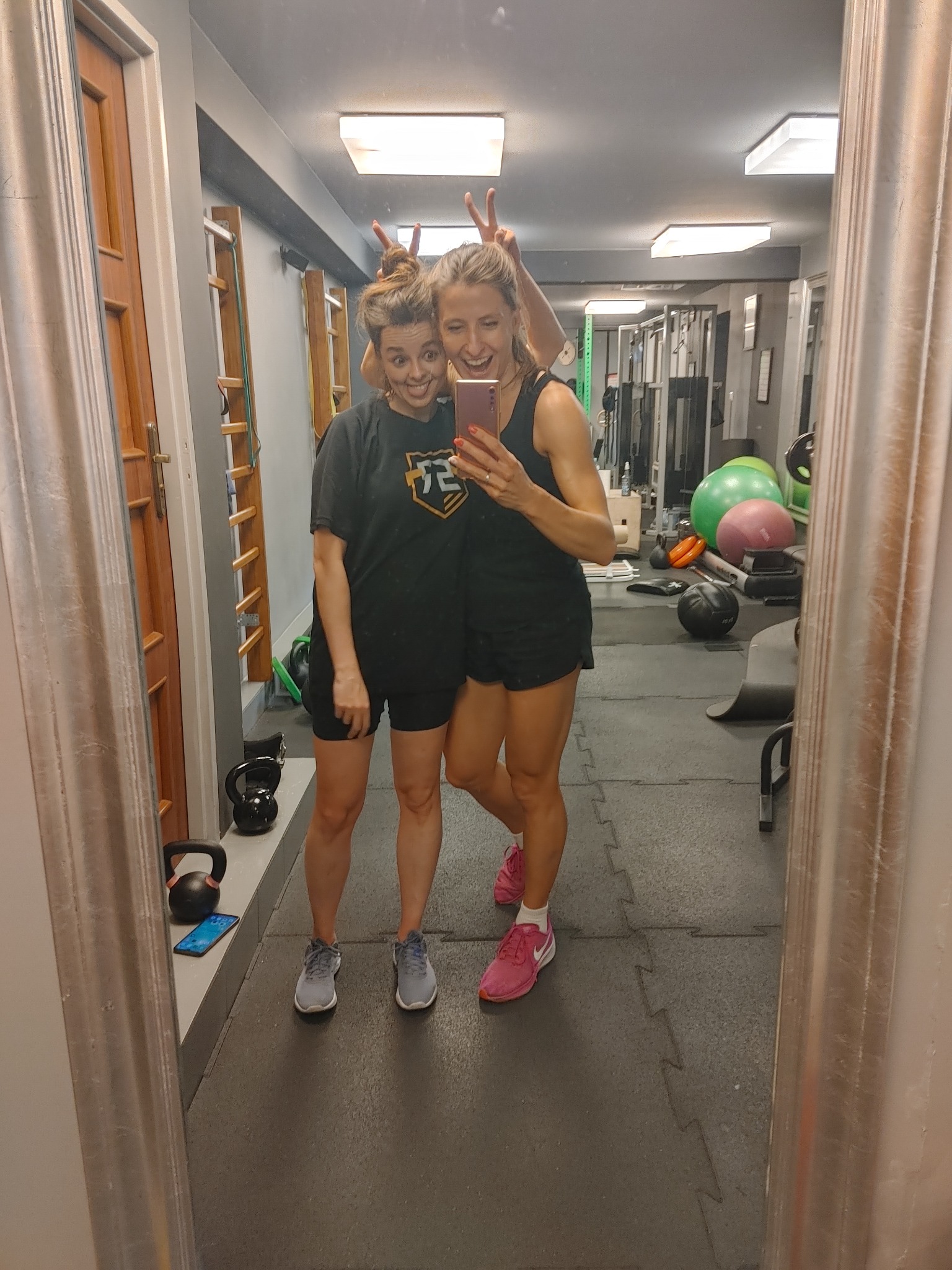 Dwie uśmiechnięte kobiety robią selfie w lustrze w siłowni, jedna z nich robi uszy królika za głową drugiej, w tle drabinki gimnastyczne i sprzęt do ćwiczeń.