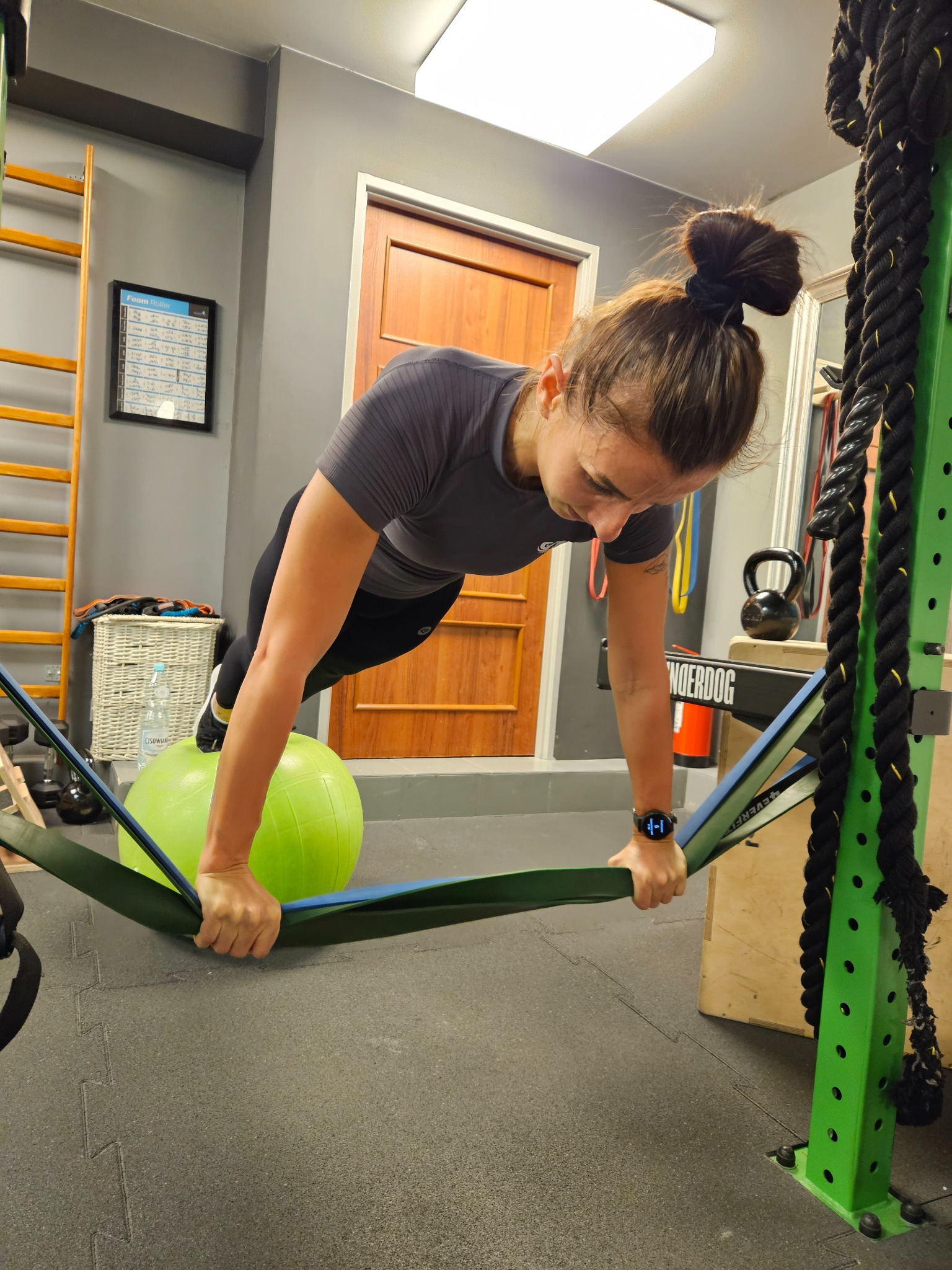 Kobieta w szarym topie i czarnych legginsach wykonuje pompkę na zielonej piłce gimnastycznej, trzymając elastyczne taśmy treningowe w dłoniach, w tle drabinka gimnastyczna, kettlebell i liny...