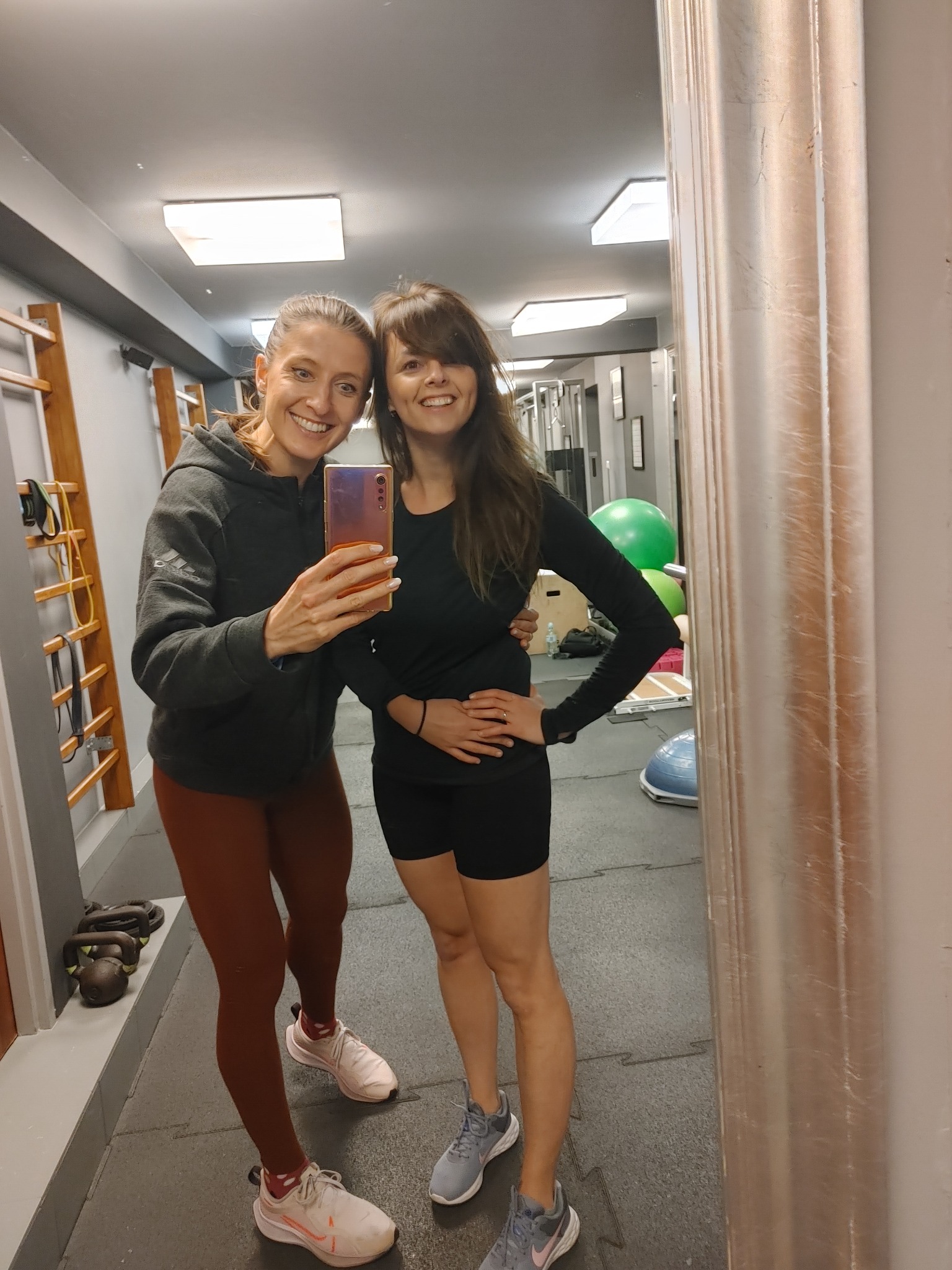 Dwie uśmiechnięte kobiety w strojach sportowych robią selfie w siłowni; w tle drabinki gimnastyczne, kettlebells i piłki do ćwiczeń.