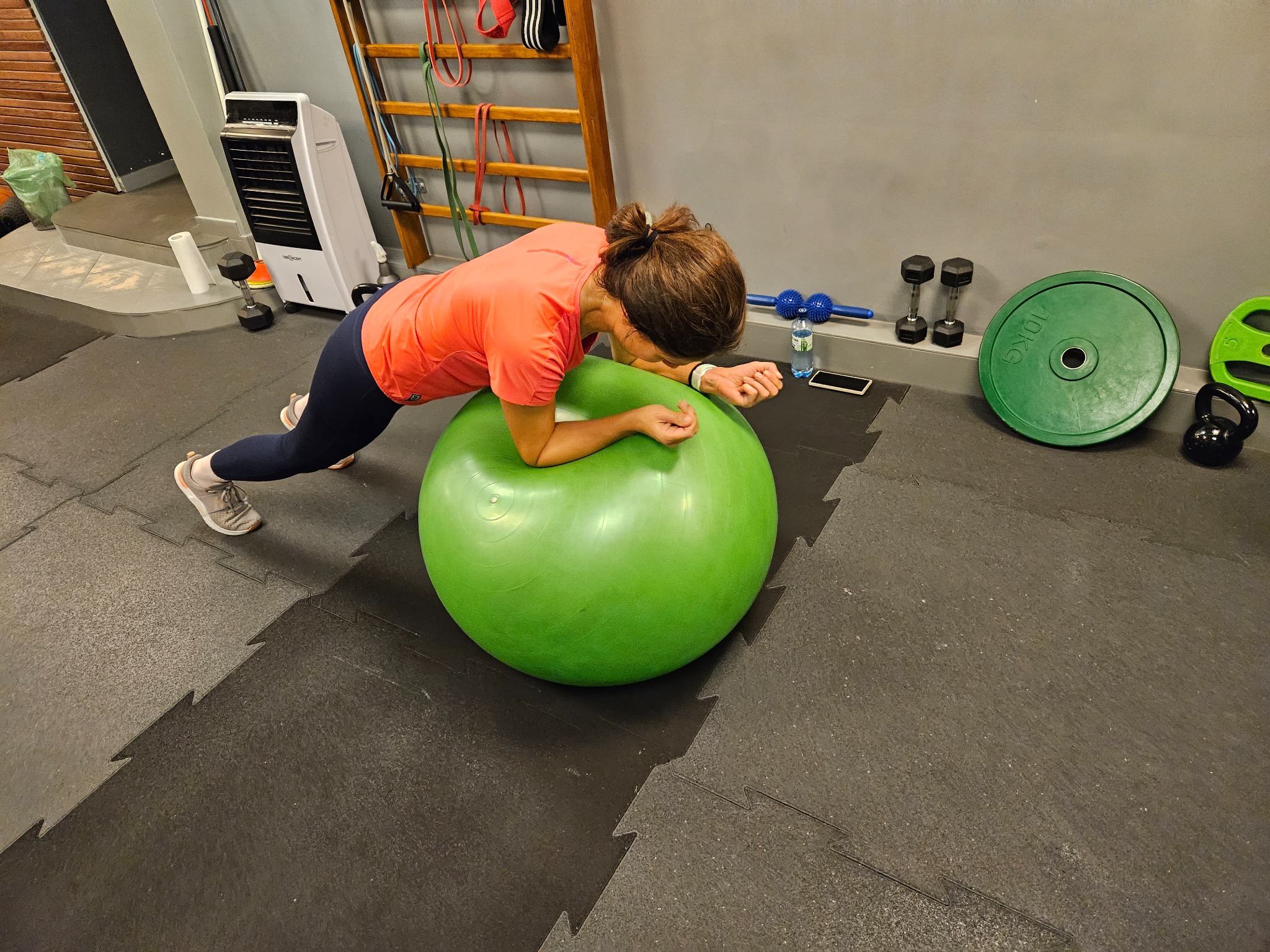 Kobieta w pozycji plank na zielonej piłce gimnastycznej w siłowni, widoczne ciężarki, mata podłogowa, drabinka i nawilżacz powietrza.
