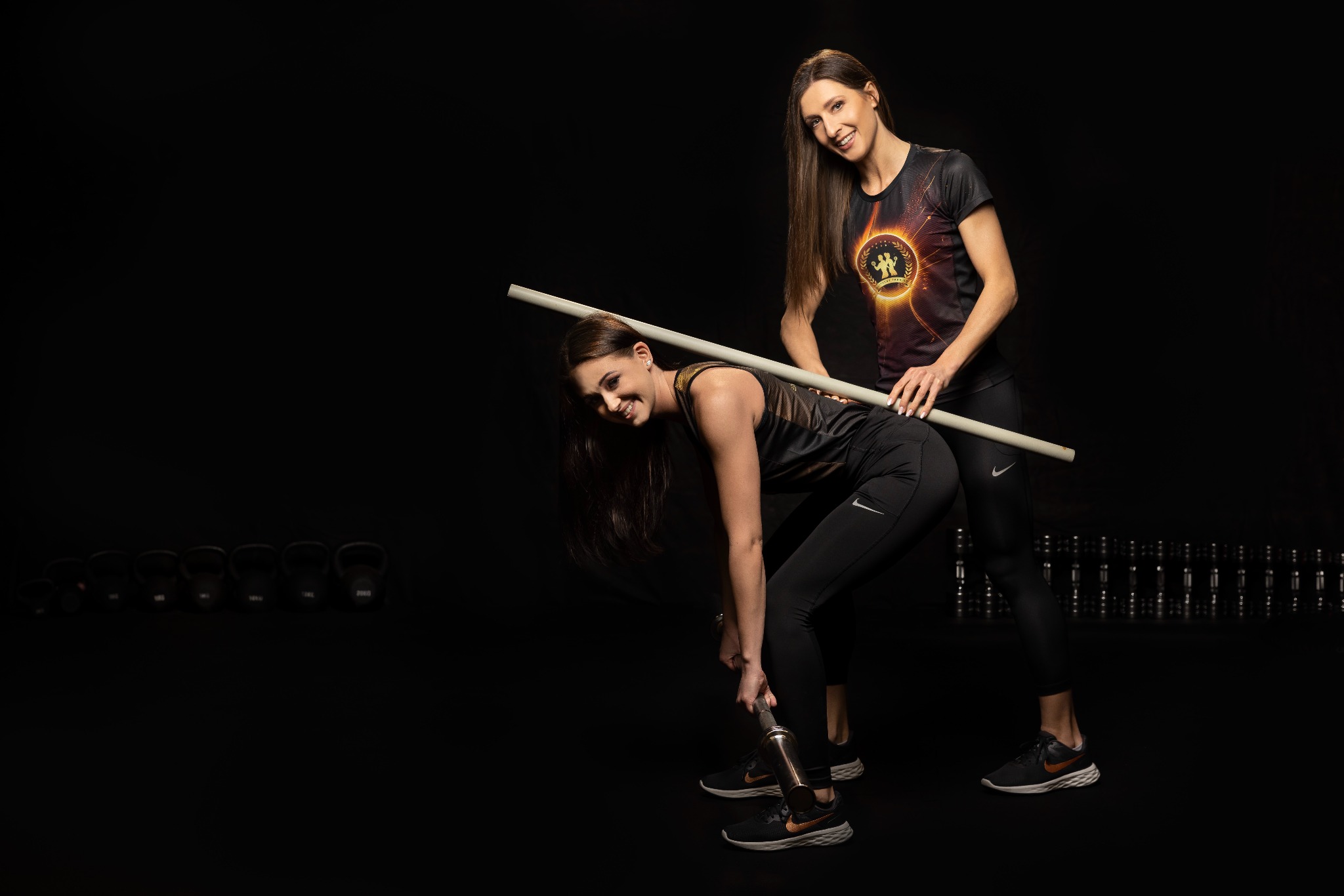 Kobieta w czarnym stroju sportowym wykonuje ćwiczenie ze sztangą pod nadzorem instruktorki z Tarnowca, w tle widoczne kettlebells.