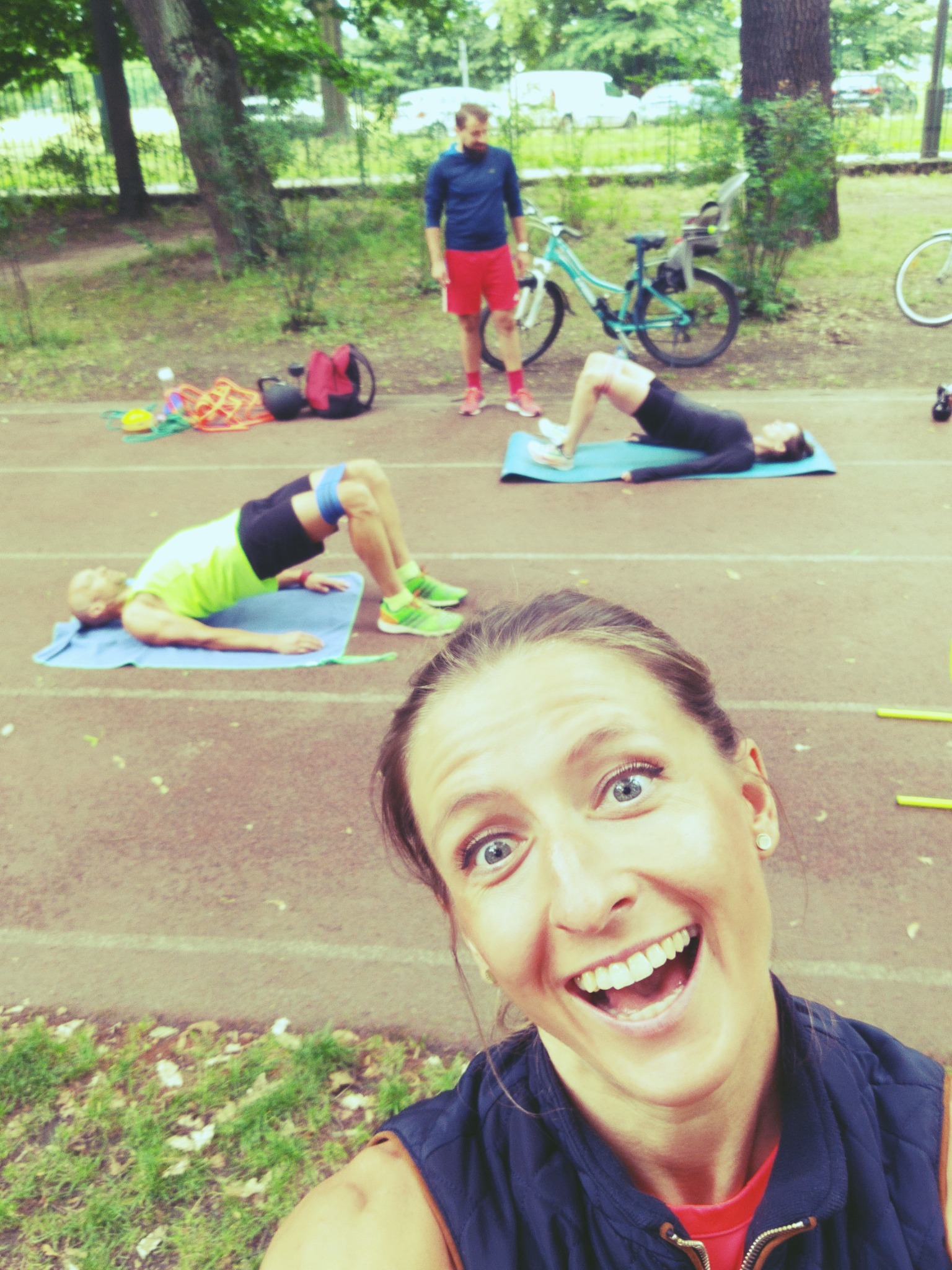 Uśmiechnięta kobieta robi selfie na bieżni, w tle dwie osoby ćwiczące unoszenie bioder na matach, mężczyzna w czerwonych spodenkach i rower.