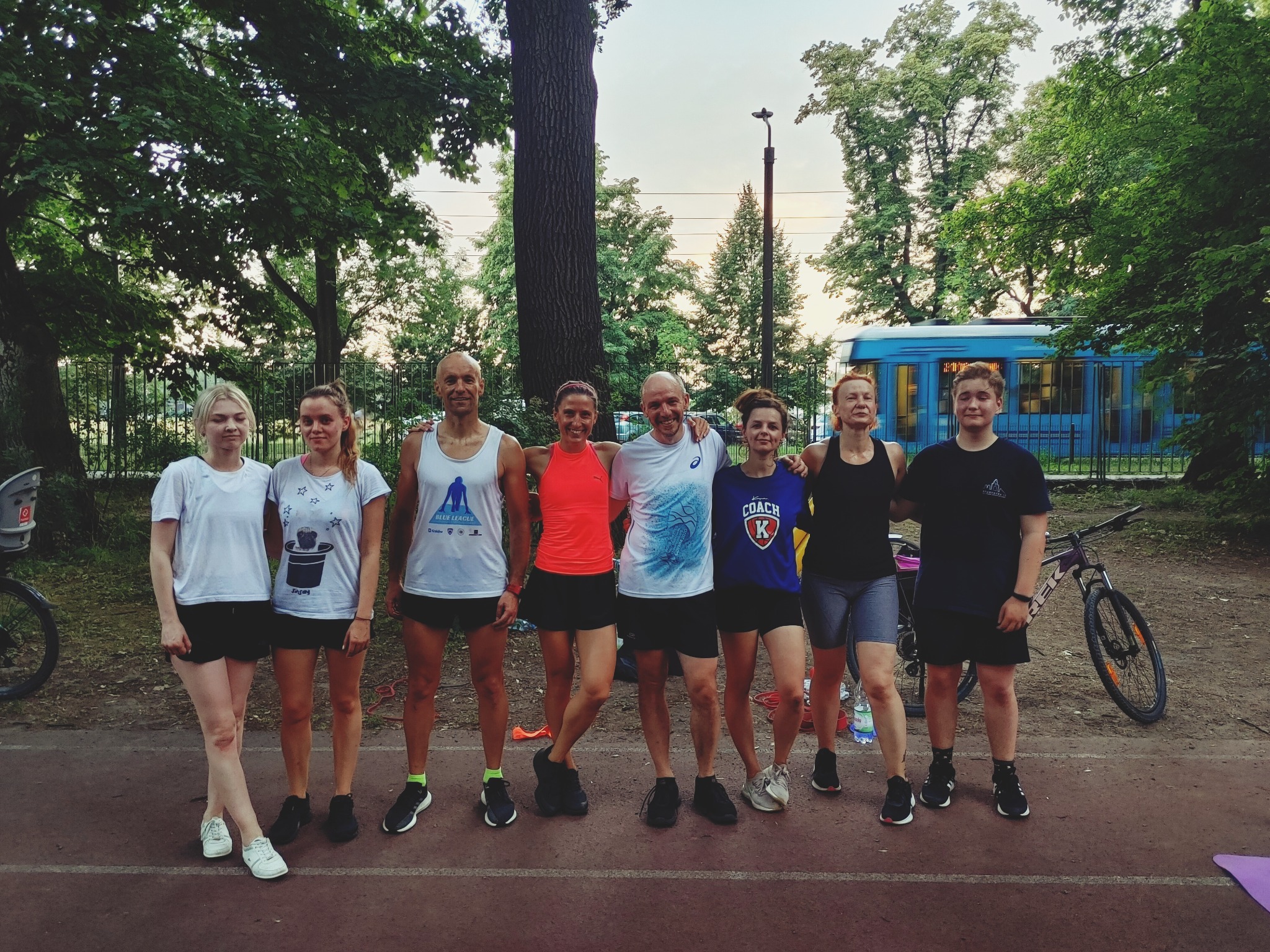 Grupa ośmiu osób w strojach sportowych, stojących na bieżni w parku, z tramwajem w tle, pozujących do zdjęcia po treningu.