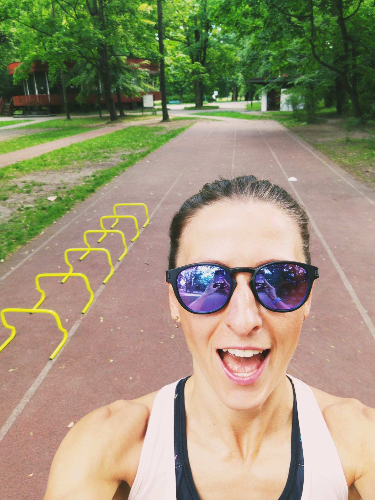 Uśmiechnięta kobieta w okularach przeciwsłonecznych robiąca selfie na bieżni z żółtymi płotkami do ćwiczeń w tle.