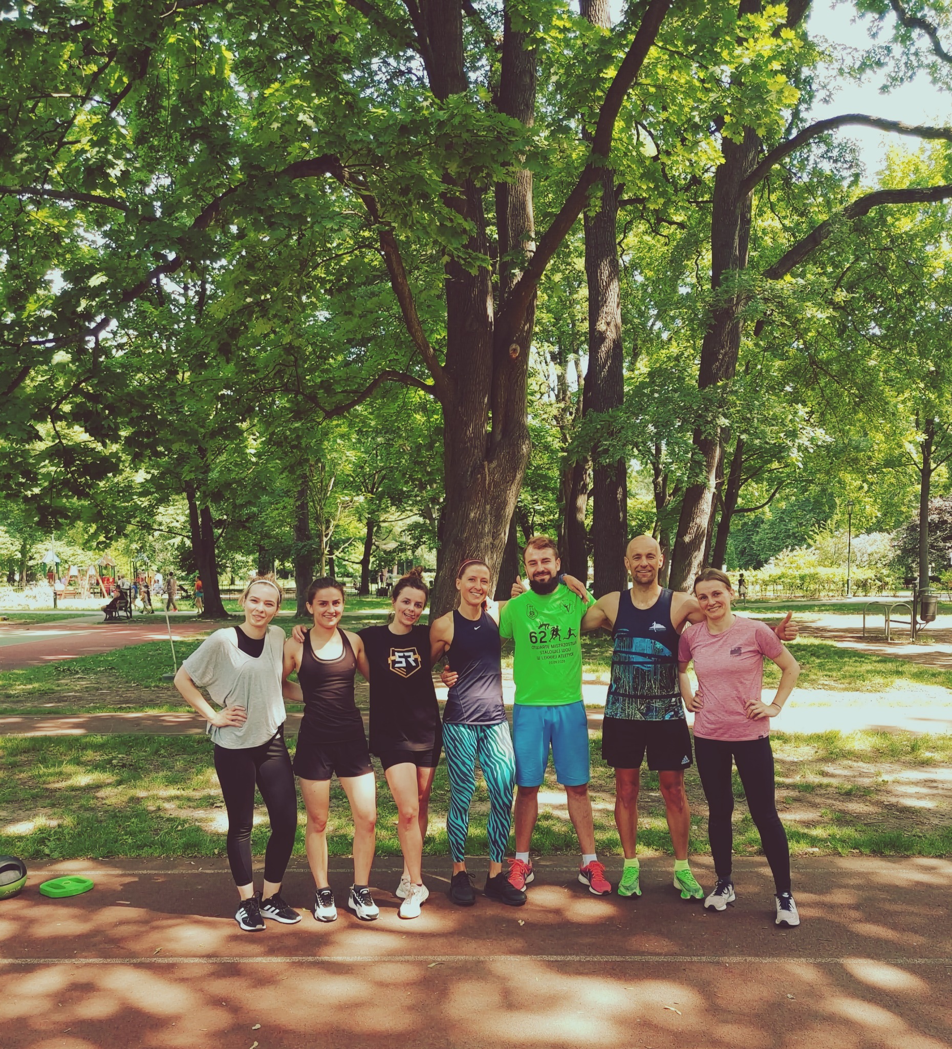 Grupa sześciu osób w strojach sportowych pozuje na tle parku, drzewa i w tle widoczny plac zabaw.