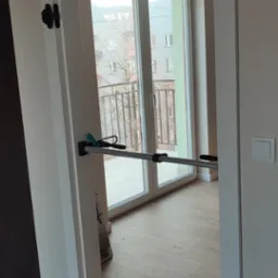 Biała ościeżnica drzwiowa tymczasowo ustabilizowana rozpórkami teleskopowymi podczas montażu, widok na balkon i parkiet