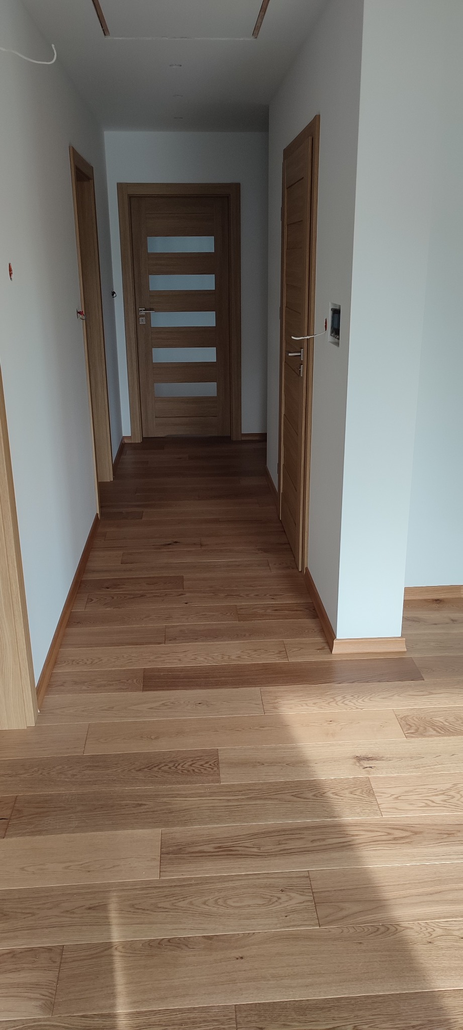 Korytarz z nową podłogą z jasnych paneli drewnianych i dwiema zamontowanymi drzwiami z naturalnego drewna, jedna z przeszkleniami.