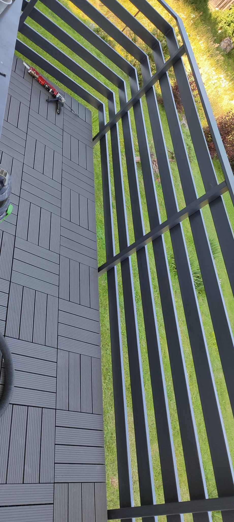 Montaż szarych, kompozytowych płytek podłogowych na tarasie, widoczna fuga, narzędzie do klejenia i metalowa konstrukcja pergoli nad tarasem, widok z góry na trawnik.