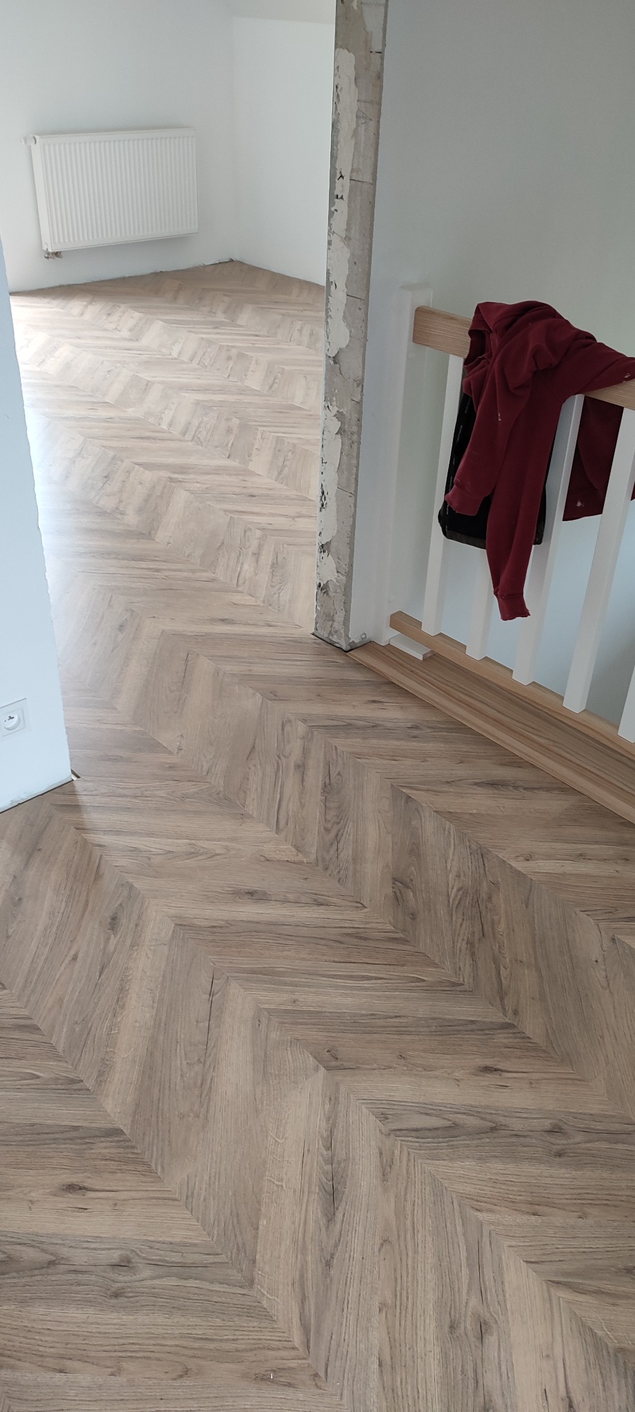 Ułożona pod kątem jodełki podłoga z paneli w odcieniach brązu, widoczne fragmenty schodów z białą balustradą i niedokończone wykończenie słupa.