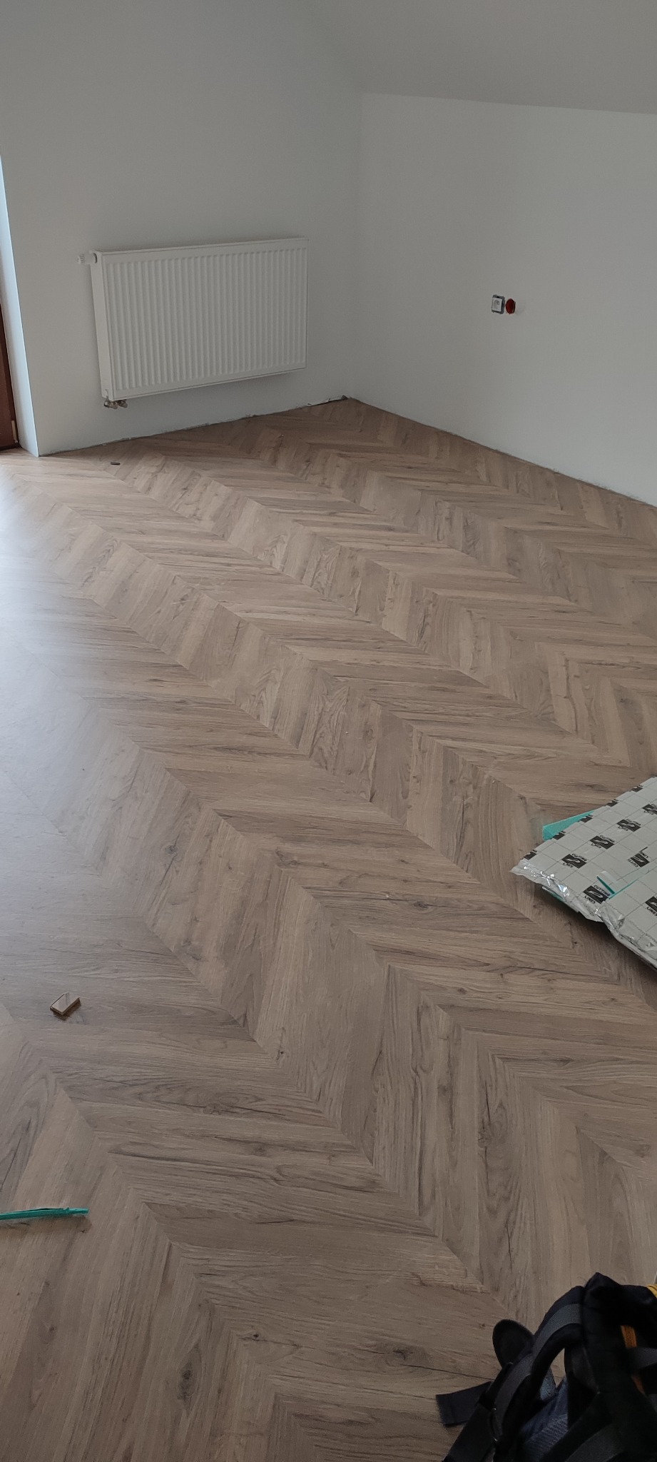 Ukończone układanie parkietu w jodełkę francuską, widoczny grzejnik na ścianie, fragment podkładu pod panele i torba narzędziowa.