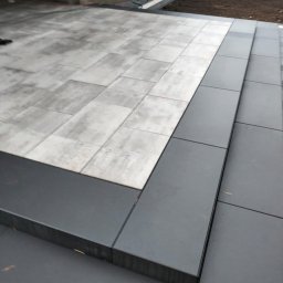 Kolo-Bruk - Ułożona nawierzchnia z jasnoszarej kostki brukowej, z ciemnoszarymi obrzeżami i schodami, prowadząca do świeżo przygotowanego klombu z ziemią.
