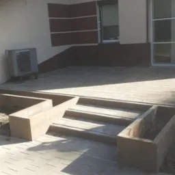 Ułożona kostka brukowa prowadząca do domu z trzema stopniami i betonowymi donicami po bokach, widoczny klimatyzator na ścianie budynku.