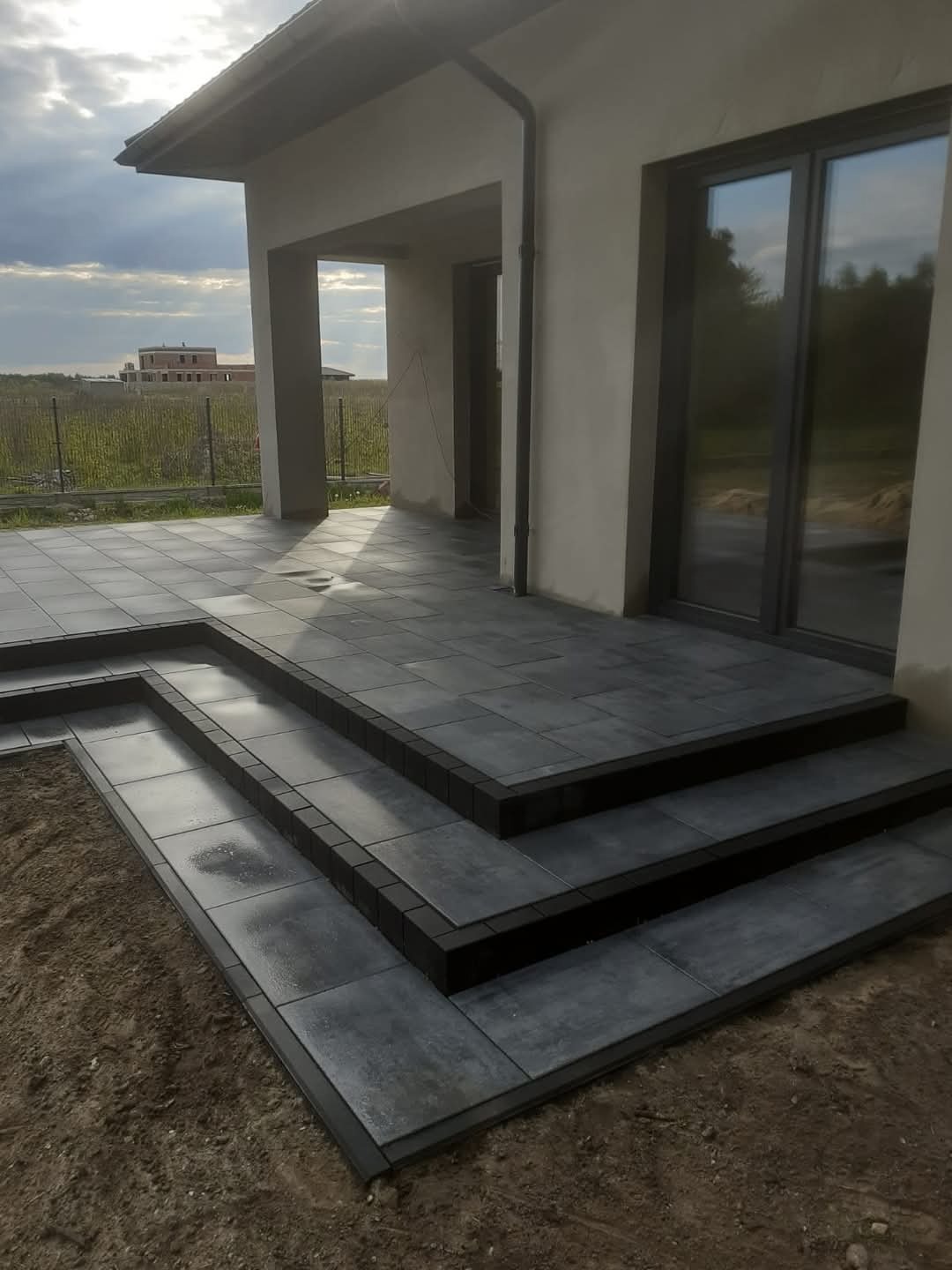 Nowoczesny taras z szarych płyt betonowych i schodami, z widokiem na budynek w tle i zachmurzone niebo.