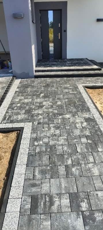 Ułożona szara kostka brukowa prowadząca do wejścia domu, z obramowaniem z jasnej kostki granitowej i fragmentem przygotowanego podłoża po bokach.