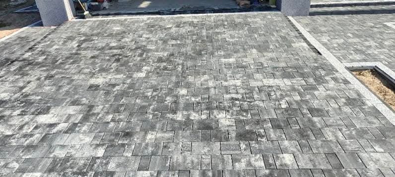 Ułożona szara kostka brukowa w geometryczny wzór, obrzeża z jasnoszarego granitu, widoczne słupy wejściowe w tle.