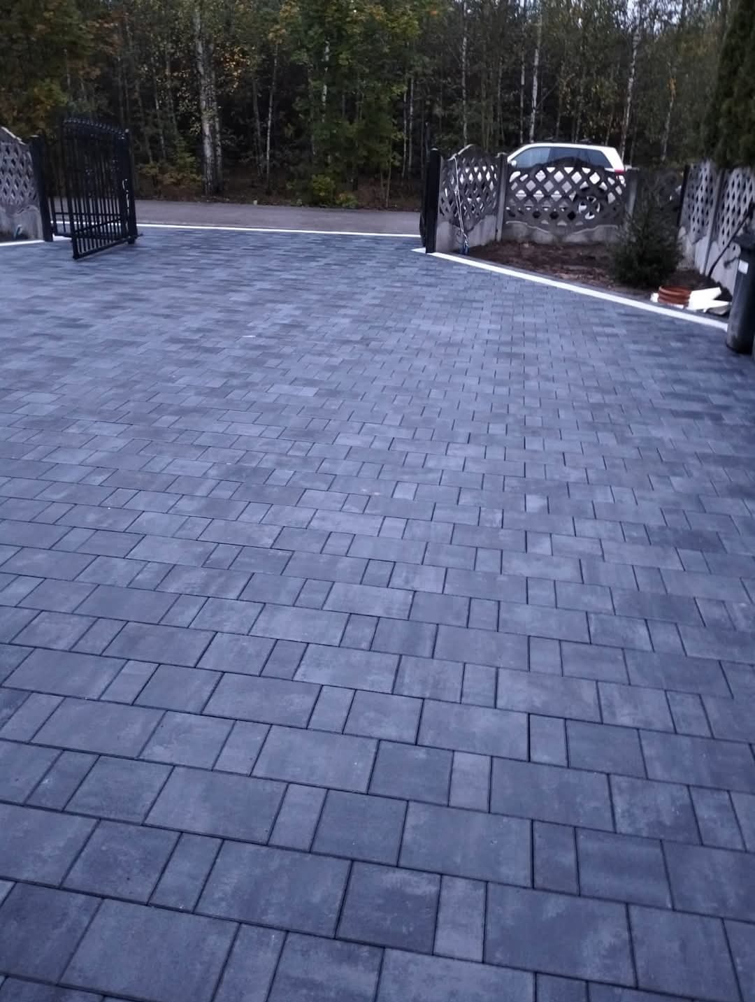 Ułożona, grafitowa nawierzchnia z kostki brukowej na posesji, z widoczną bramą wjazdową i fragmentem ogrodzenia z betonowych prefabrykatów. W tle drzewa i zaparkowany samochód.