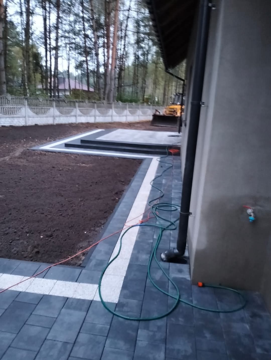 Ułożona szara kostka brukowa z białym obramowaniem, prowadząca do betonowych schodów przed domem. W tle widoczny trawnik, ogrodzenie panelowe i koparka.
