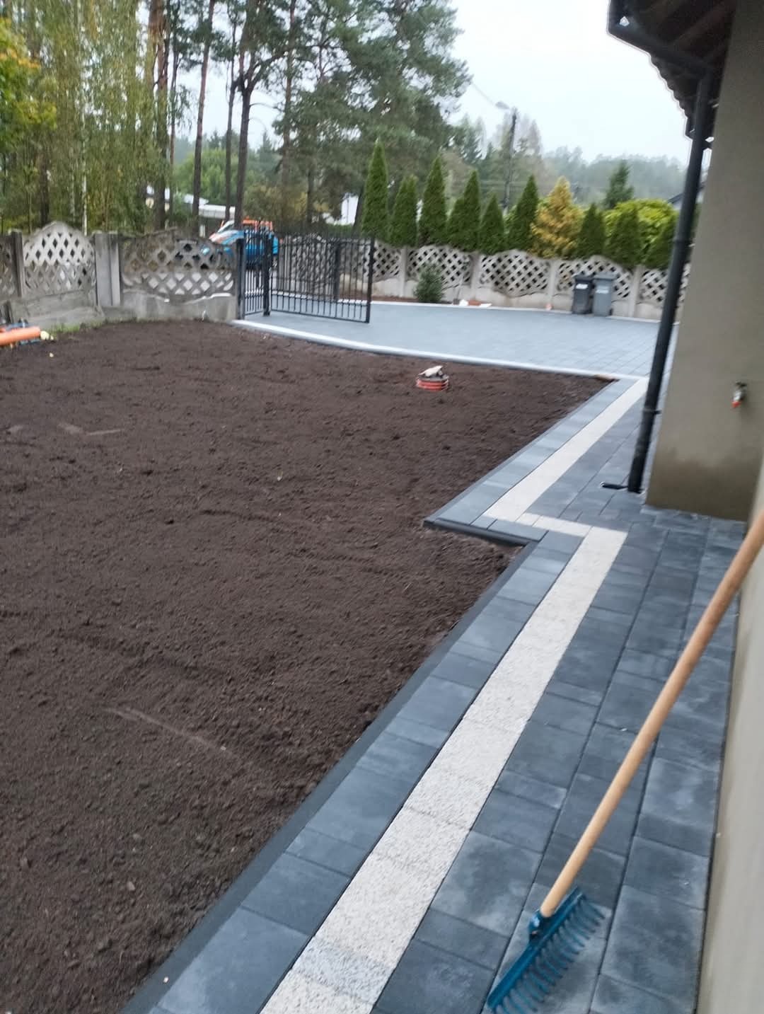 Przygotowany teren pod trawnik z widoczną świeżą ziemią, obok ułożona kostka brukowa z jasnym pasem, grabie opierają się o ścianę budynku.