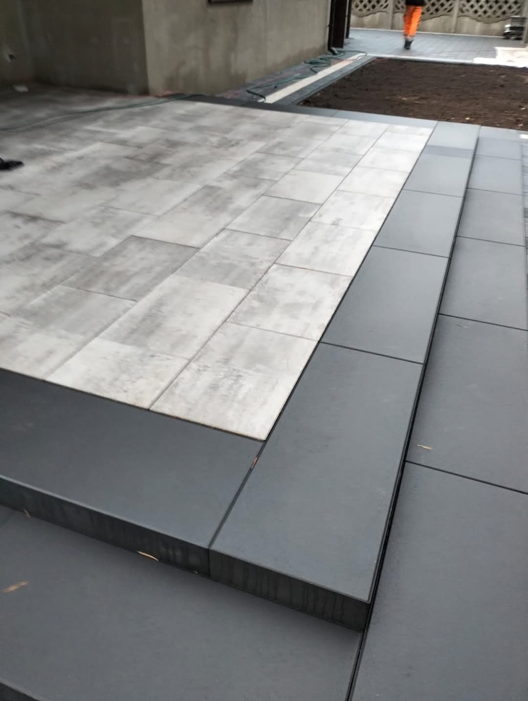 Ułożona nawierzchnia z jasnoszarej kostki brukowej, z ciemnoszarymi obrzeżami i schodami, prowadząca do świeżo przygotowanego klombu z ziemią.