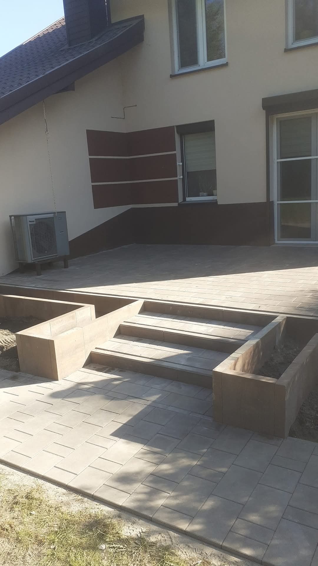 Ułożona kostka brukowa prowadząca do domu z trzema stopniami i betonowymi donicami po bokach, widoczny klimatyzator na ścianie budynku.