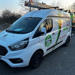 Łukasz Janus - Biały van z logo firmy czyszczącej ciśnieniowo, zaparkowany na kostce brukowej. Na dachu drabina. Widoczne hasła: 'Mycie ciśnieniowe bruk elewacja dachy'.