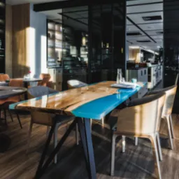 Stół z blatem z drewna i żywicy epoksydowej w kolorze turkusowym, ustawiony w restauracji, widoczne krzesła z drewnianymi oparciami i szarą tapicerką, na blacie leży książka i szklana karafka z wodą.