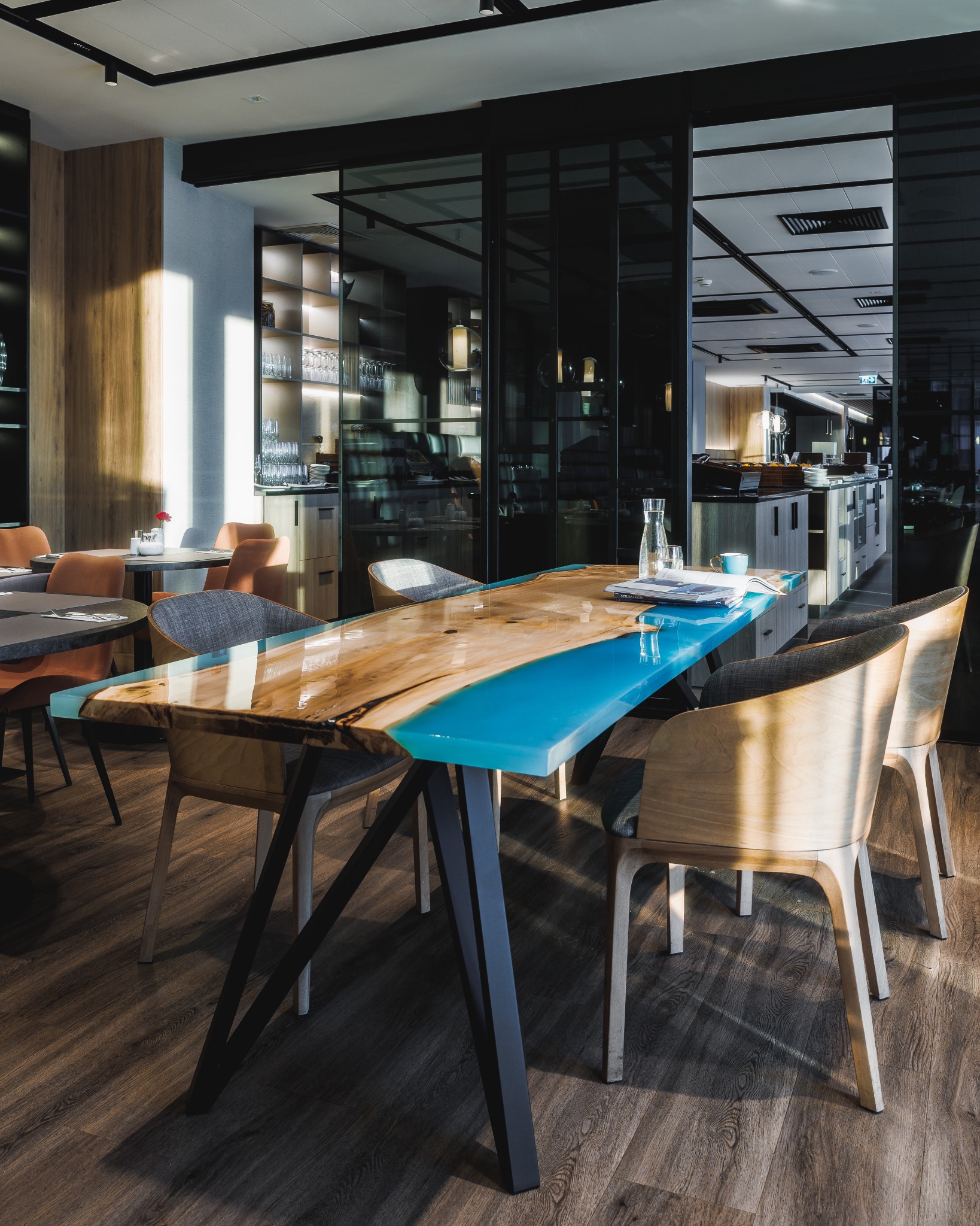 Stół z blatem z drewna i żywicy epoksydowej w kolorze turkusowym, ustawiony w restauracji, widoczne krzesła z drewnianymi oparciami i szarą tapicerką, na blacie leży książka i szklana karafka z wodą.
