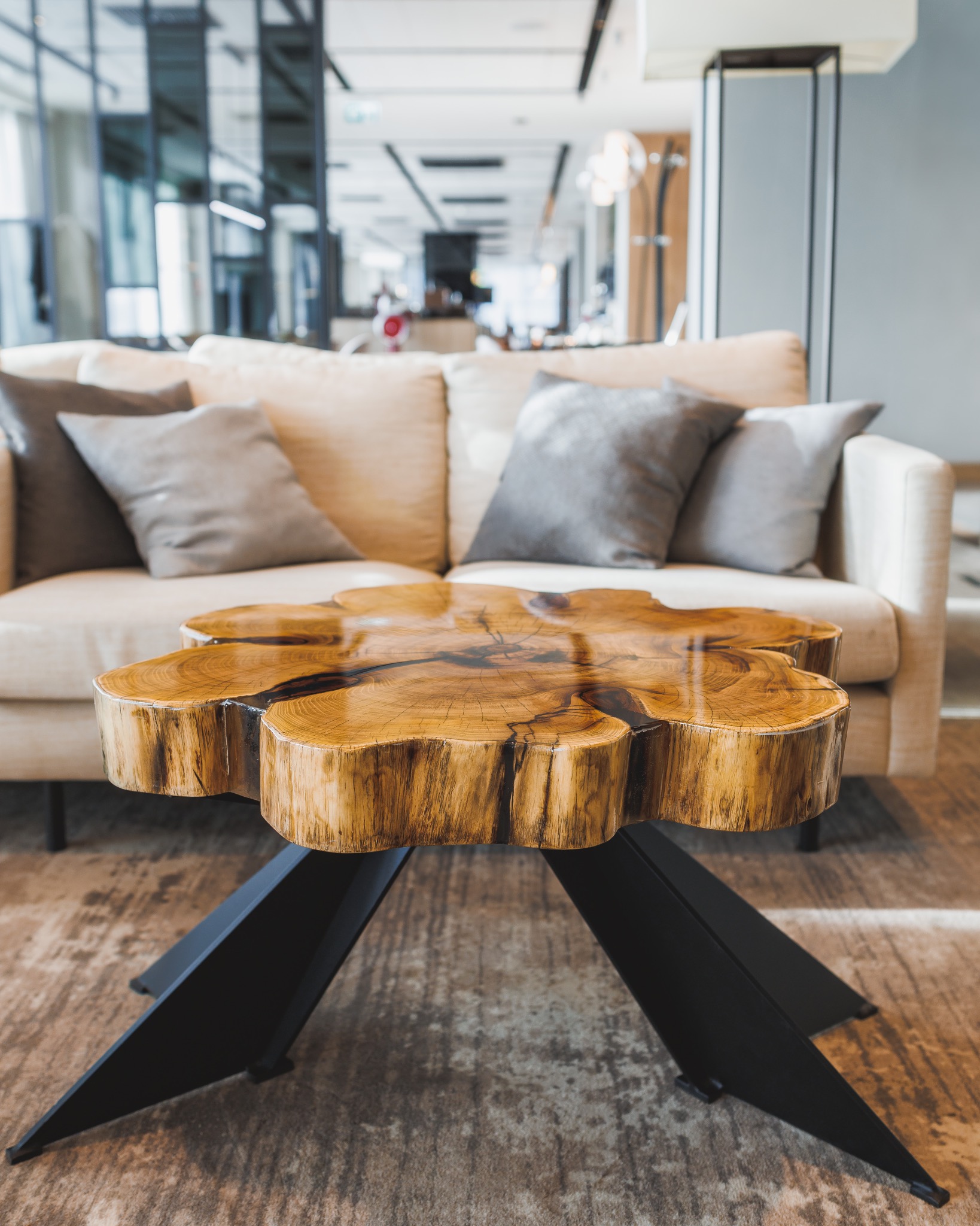 Stolik kawowy z blatem z plastra drewna o nieregularnym kształcie i czarnych, metalowych nogach na tle beżowej sofy z poduszkami w nowoczesnym wnętrzu.