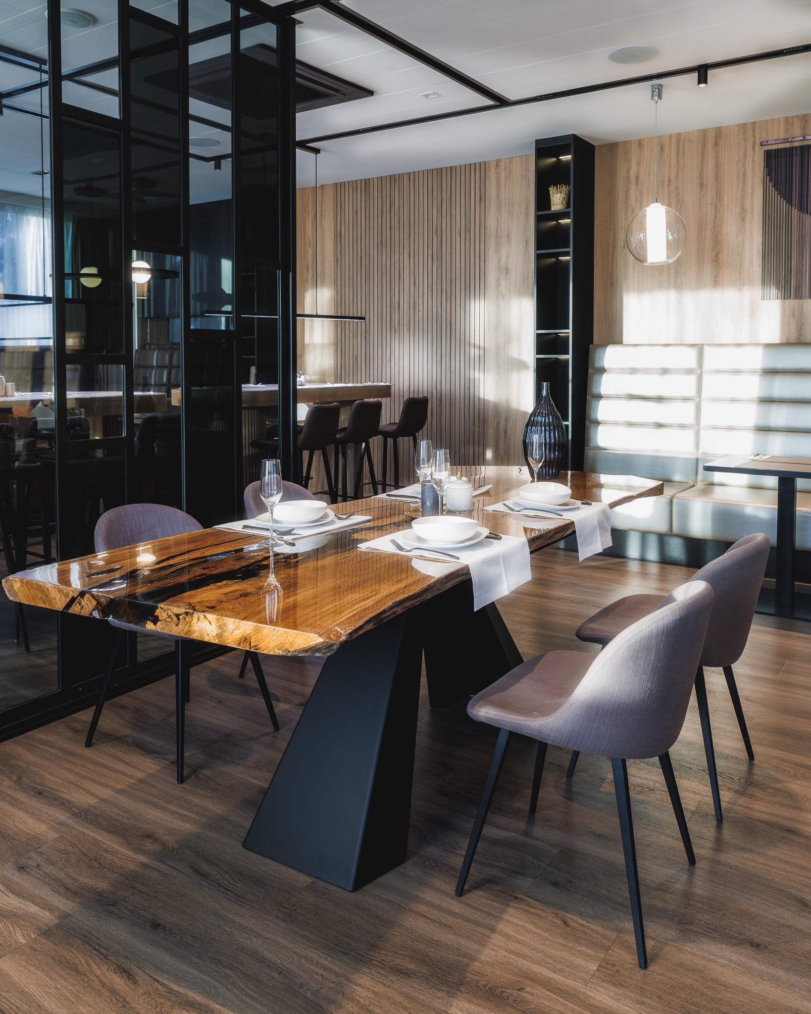 Jadalnia z blatem z żywicy epoksydowej i drewna, czarne, trapezowe nogi stołu, szare krzesła, nakryty stół, drewniana boazeria ścienna w tle.