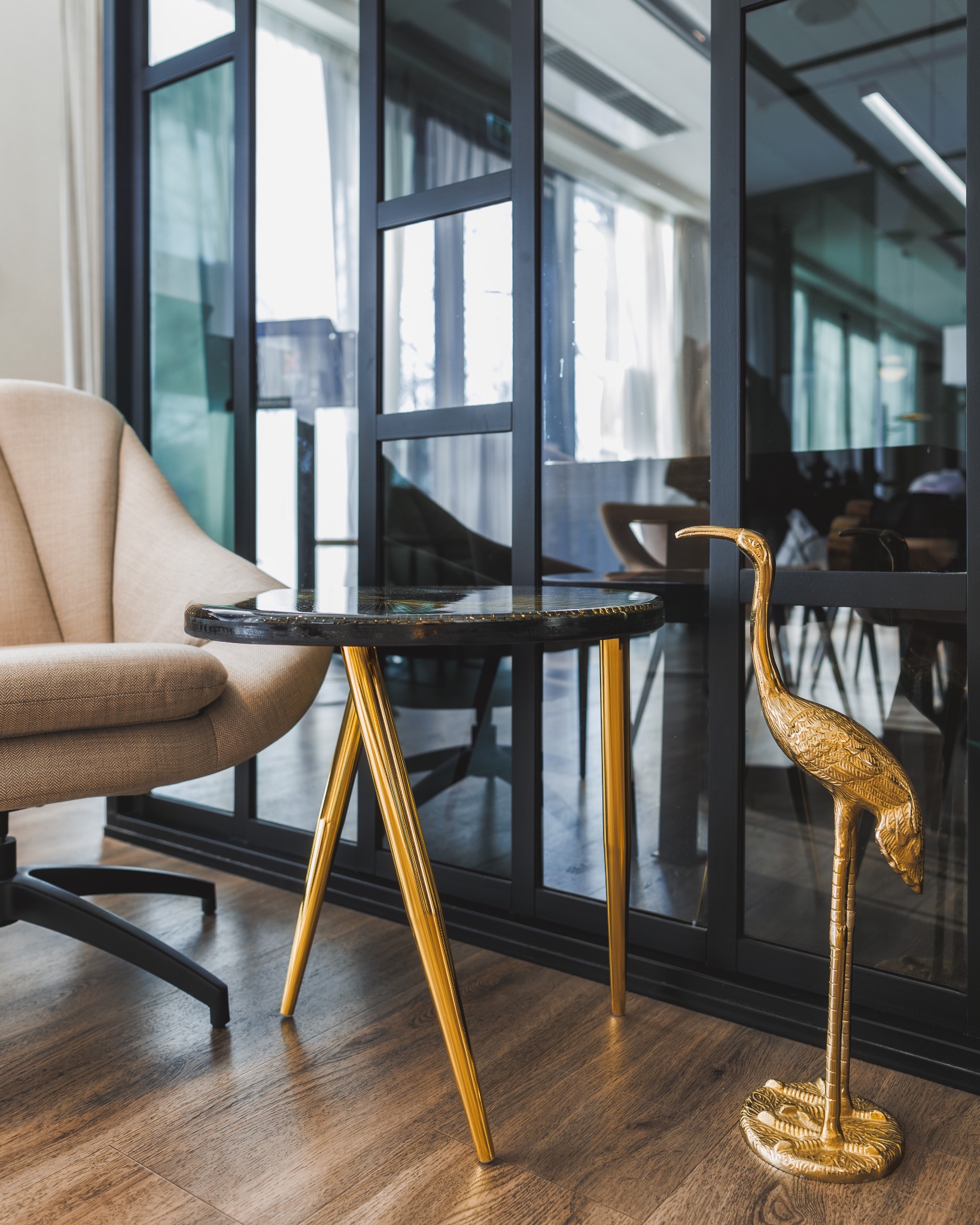 Stylowy blat okrągłego stolika kawowego z czarnym blatem i złotymi nogami, obok fotela i złotej figurki żurawia na drewnianej podłodze.