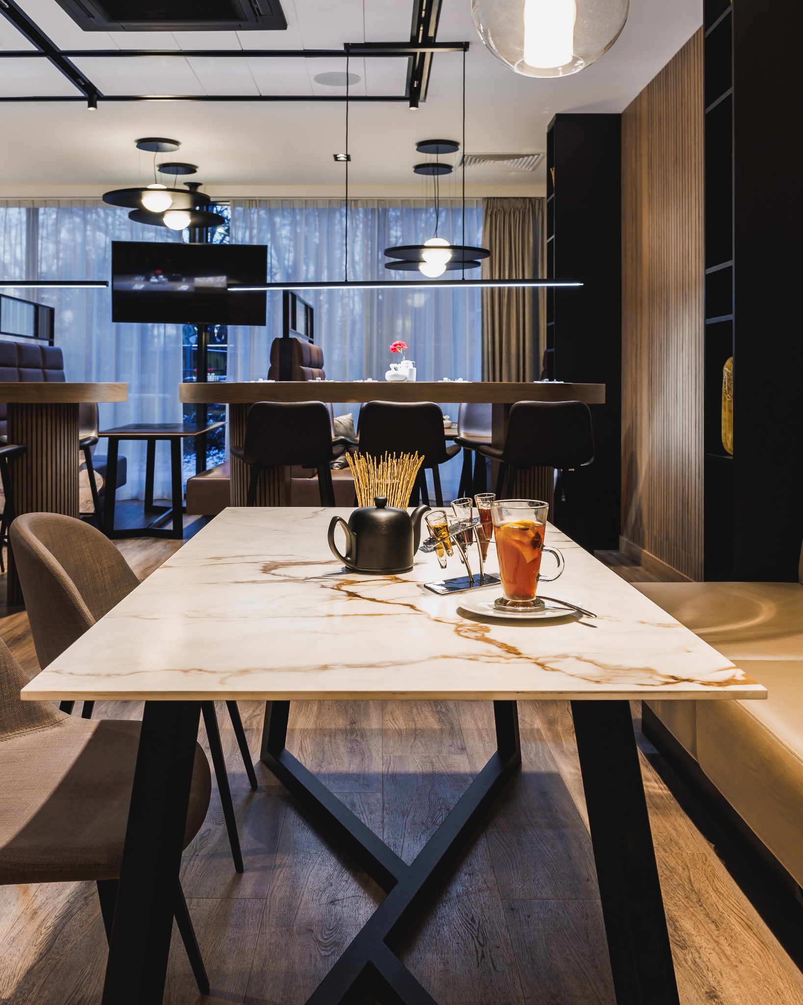 Marmurowy blat stołu w kawiarni z czarną podstawą, na nim czajnik, szklanka herbaty i słomki, w tle inne stoliki i krzesła.