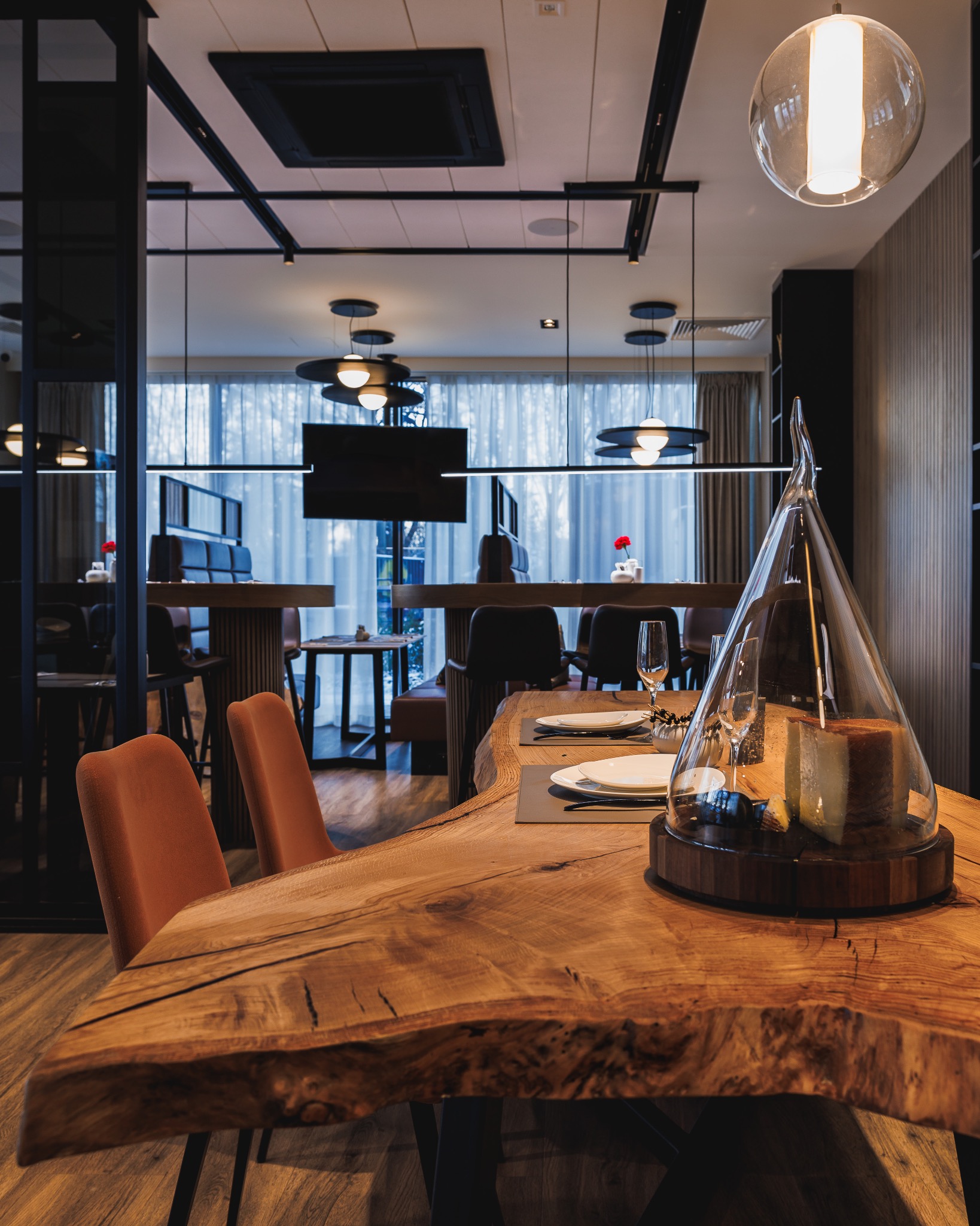 Rustykalny blat stołu z litego drewna o nieregularnych krawędziach, nakryty dwoma zestawami talerzy i szklanek, z serami pod szklanym kloszem, w nowoczesnym wnętrzu restauracji.