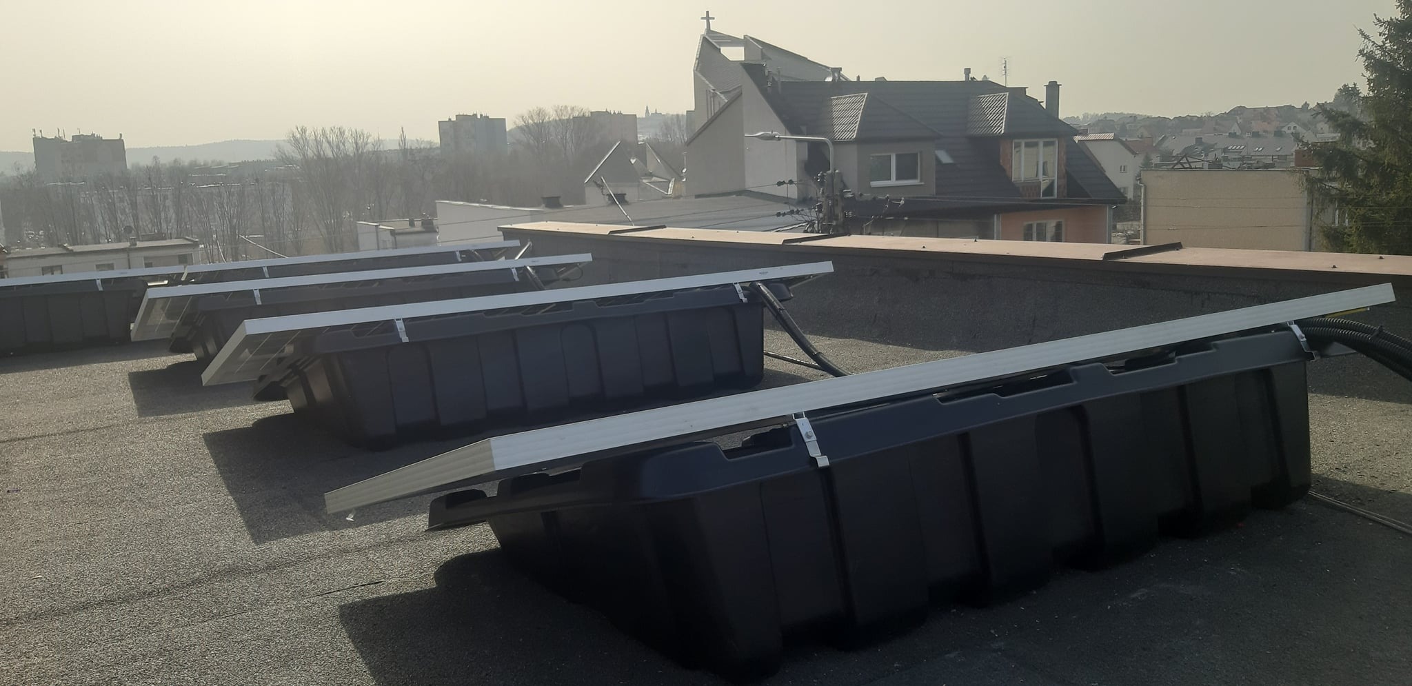 Rząd czarnych, plastikowych pojemników balastowych z panelami słonecznymi na płaskim dachu z widokiem na miasto i kościół w tle, ujęcie w pochmurny dzień.