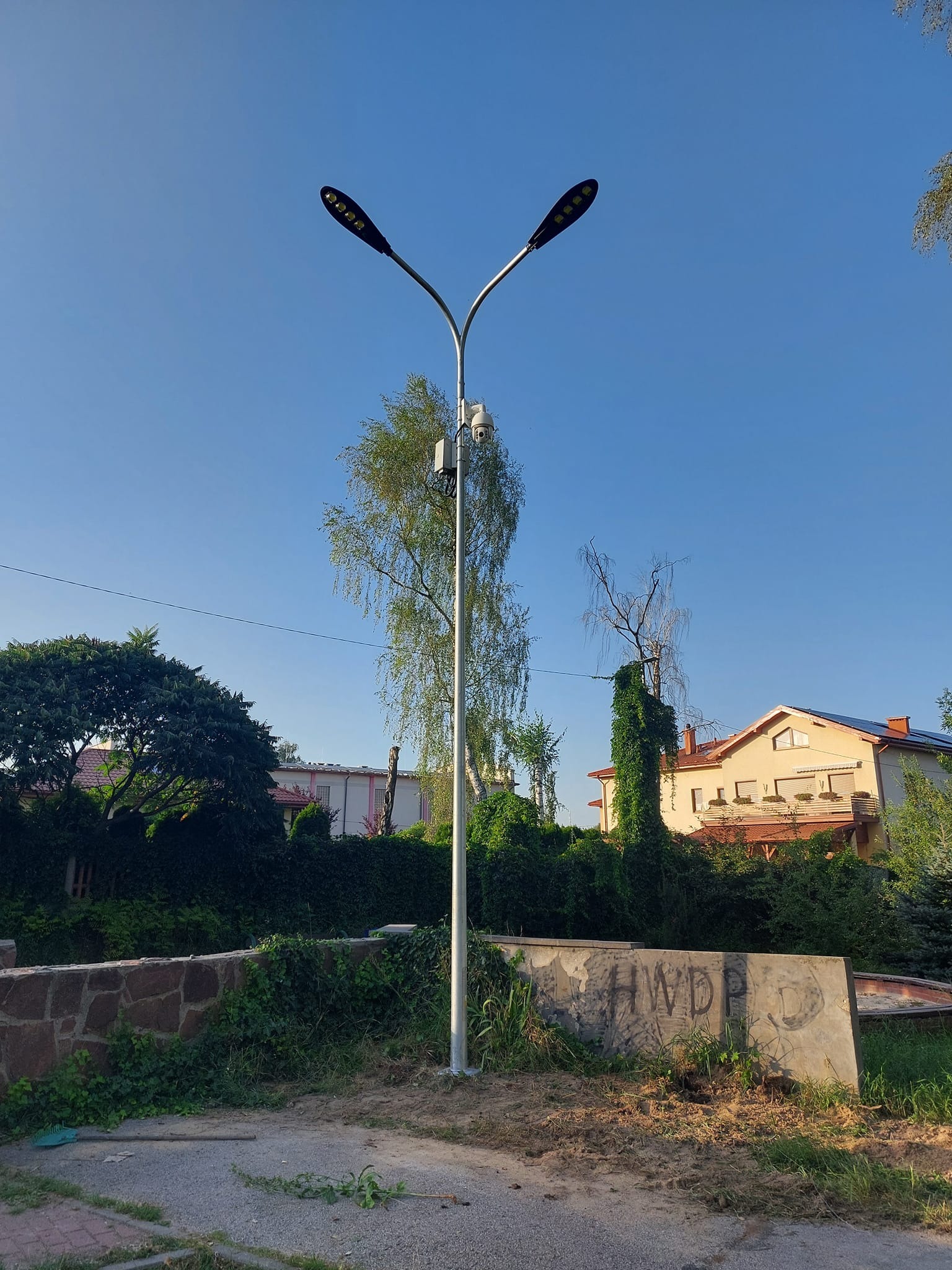 Słup oświetleniowy LED z zamontowaną kamerą monitoringu na tle błękitnego nieba, otoczony zielenią i zabudową jednorodzinną.