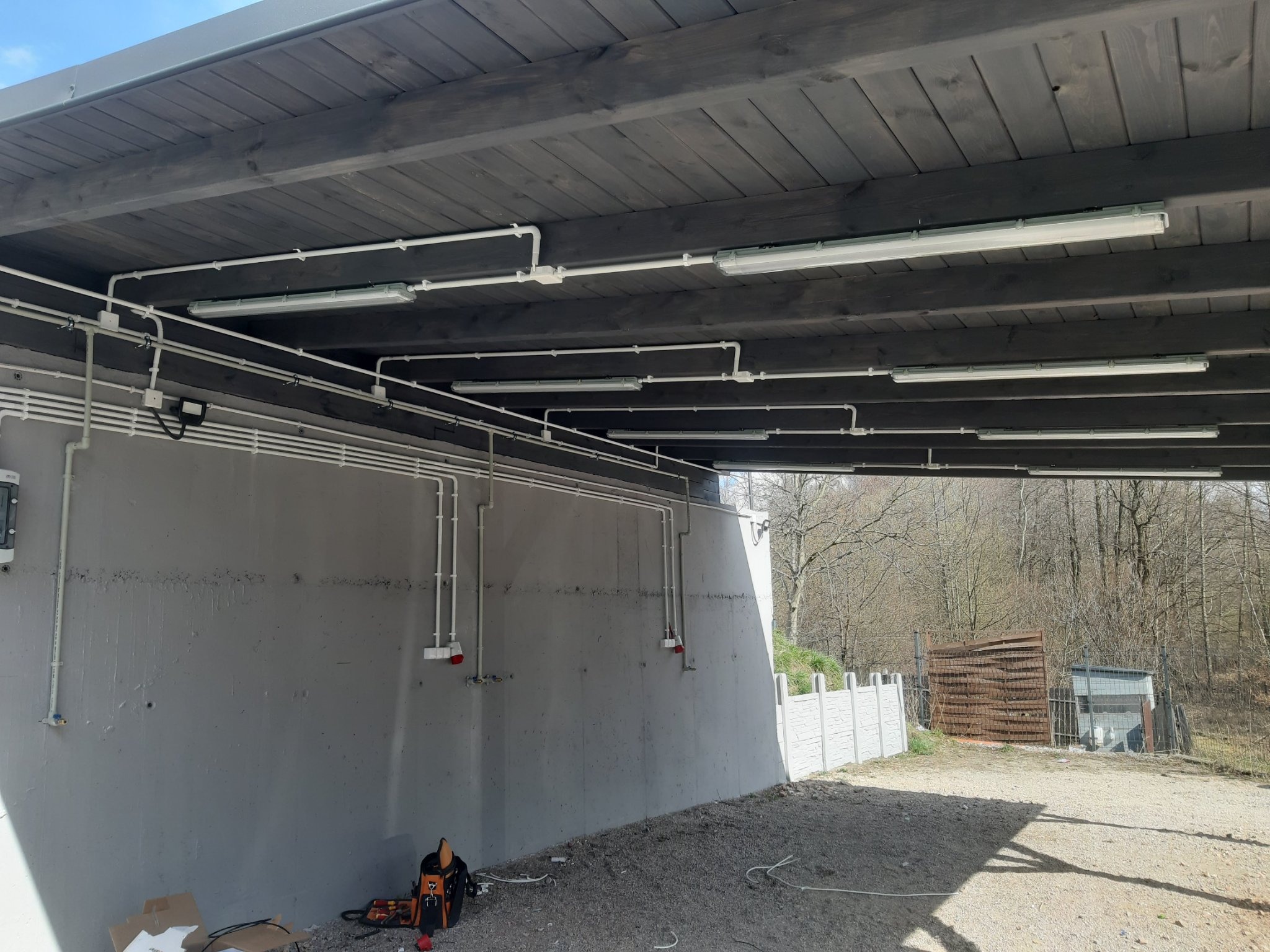 Instalacja elektryczna w garażu podziemnym: białe rury i lampy fluorescencyjne zamontowane na ciemnoszarym, drewnianym suficie, z widocznymi narzędziami i skrzynką elektryczną na szarej ścianie.