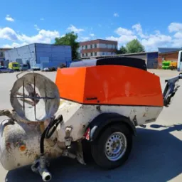 Agregat tynkarski z pomarańczową obudową i mieszadłem, zaparkowany na placu budowy, widoczny wózek widłowy w tle.