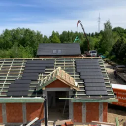 Montaż czarnej dachówki ceramicznej na nowym budynku, widoczna konstrukcja więźby dachowej i częściowo ułożone pokrycie, w tle dźwig budowlany i zieleń drzew.