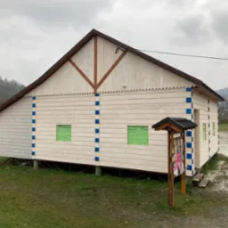 Nowa drewniana konstrukcja budynku z widoczną więźbą dachową, obita jasnymi deskami z niebieskimi akcentami, na podwyższeniu, z widocznymi oknami zabezpieczonymi folią i słupem ogłoszeniowym obok...