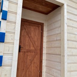 Fox House - Wejście do drewnianej altany z brązowymi drzwiami z ukośnym wzorem i czarną klamką, ściany w jasnym odcieniu drewna z niebieskimi akcentami, widok z zewnątrz.