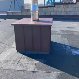 Brązowa obudowa komina z metalowym zakończeniem na dachu pokrytym papą, w tle fragment parku rozrywki z kolejką górską.