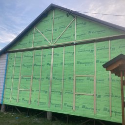 Fox House - Budynek w trakcie ocieplania, widoczna zielona membrana Eurovent i drewniane listwy pionowe, fragment ściany obłożony sidingiem.